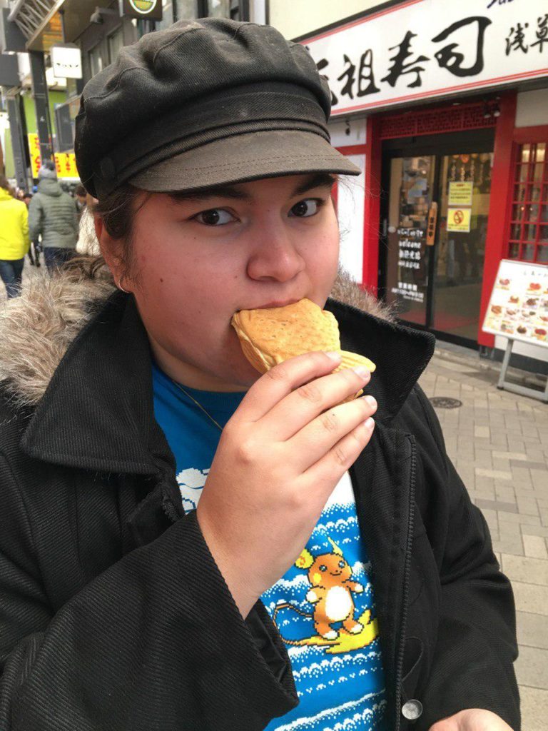 A photo of Chu eating taiyaki on a street in Japan. They are wearing a black hat, a back coat, and a blue shirt with the Pokemon Raichu on it.
